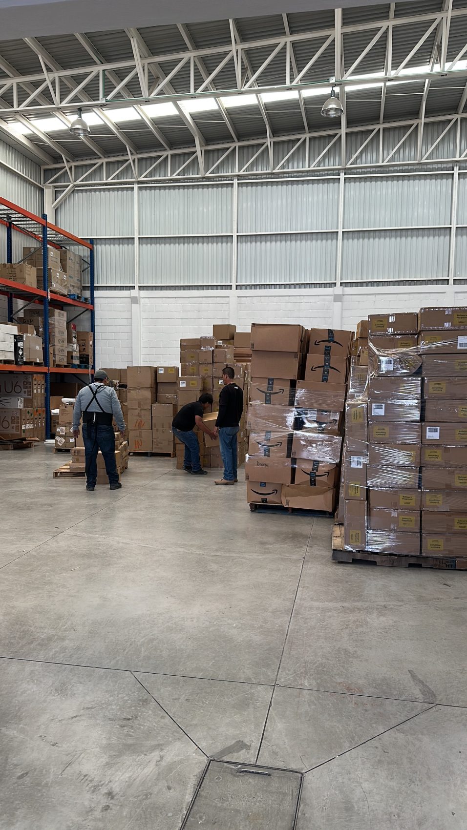 Interior de la bodega de MYYX en El Marqués: tres personas acomodando cajas sobre tarimas bajo la nave industrial con tragaluz